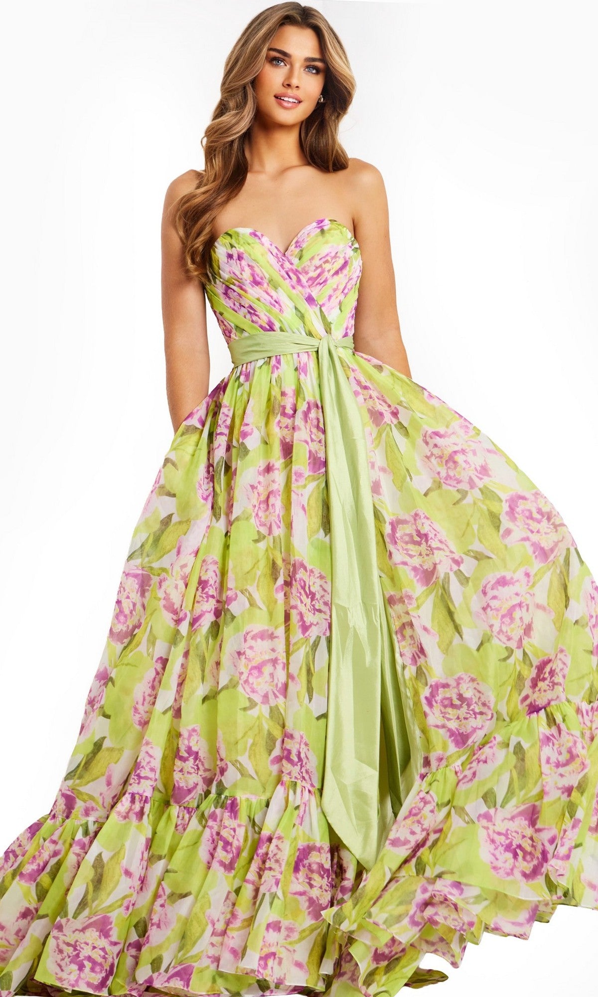 A woman wears the Jovani 48468 long prom dress in a floral print with pink and green hues, featuring a strapless, floor-length A-line silhouette and a light green sash at the waist for an elegant look.